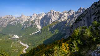 Blick auf ein sonniges Bergtal mit einem trockenen Flussbett und bewaldeten Hängen vor einer Bergkette unter blauem Himmel.