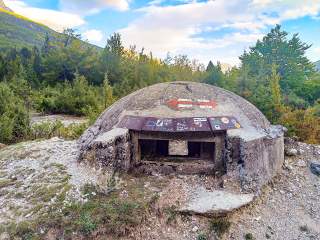 Kleiner runder Betonbunker mit Schießscharten in einer bewaldeten Berglandschaft, auf dem Dach ist ein Wanderwegzeichen mit zwei Pfeilen in entgegengesetzte Richtungen aufgemalt.