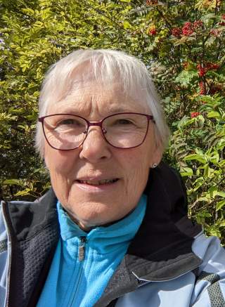 lächelnde Frau mit Brille und blauer Jacke vor Hecke