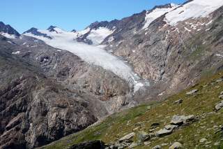 Weiße Gletscherzunge umgeben von Fels, im Hintergrund scharfe Gipfel mit Schneebedeckung