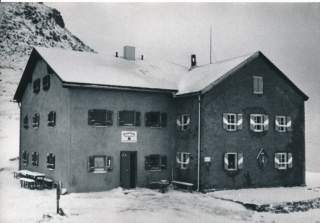 Schwarz-weiß-Foto einer großen, zweistöckigen Berghütte mit vielen Fenstern und geschlossenen Fensterläden, umgeben von Schnee in einer bergigen Winterlandschaft.