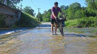 Radfahrer schiebt sein Fahrrad durch einen flachen, überschwemmten Straßenabschnitt bei Sonnenschein, umgeben von grüner Natur.