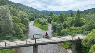 Radfahrer auf einer Brücke über einen Fluss, umgeben von dichtem Wald und Bergen.