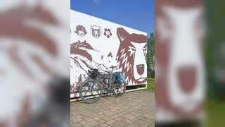 Fahrrad mit Gepäcktaschen lehnt an einer Wand mit großem Bärenmotiv und weiteren grafischen Illustrationen.
