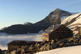 Die Neue Prager Hütte, oberhalb einer dicken Hochnebelschicht, wird von der Morgensonne beschienen.
