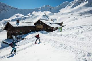 Zwei Menschen auf Skitour laufen an Berghütte vorbei