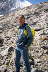 Älterer Mann in Outdoor-Bekleidung mit gelbem Rucksack steht in felsigem alpinem Gelände vor schneebedeckten Bergen.