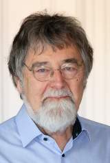 Porträt eines älteren Mannes mit grauem Haar, Bart und Brille, der ein hellblaues Hemd trägt.