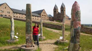 Frau in rotem Shirt betrachtet bemalte Steinsäulen auf einem Grashang, im Hintergrund ein großes historisches Klostergebäude mit zwei Türmen.