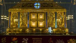 Goldener, kunstvoll verzierter Reliquienschrein mit christlichen Heiligenfiguren und Inschrift, präsentiert in einem gläsernen Schutzgehäuse.