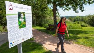Frau mit Wanderrucksack läuft an einem Informationsschild des Hildegard-Kräutergartens auf einem sonnenbeschienenen Weg entlang, umgeben von Bäumen und Grünflächen.