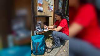 Frau in rotem Shirt kniet neben einer Stempelstation des Hildegardwegs und stempelt einen Stempelpass, neben ihr steht ein blauer Rucksack.