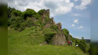 Frau in rotem Shirt kniet neben einer Stempelstation des Hildegardwegs und stempelt einen Stempelpass, neben ihr steht ein blauer Rucksack.