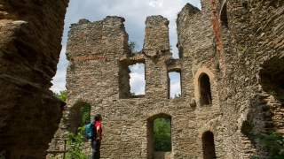 Eine Person mit Rucksack steht in einer Burgruine aus grob behauenen Steinen und blickt zu den hohen, fensterlosen Mauern, umgeben von Grün.