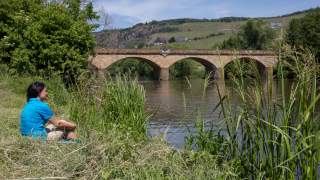 Frau sitzt am Ufer eines Flusses mit viel Grün, im Hintergrund ist eine dreibogige Steinbrücke und eine hügelige Landschaft mit Weinbergen zu sehen.
