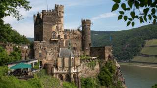 Burg auf einem Felsen mit Zinnen und runden Türmen, umgeben von grünen Weinbergen und Blick auf einen Fluss und bewaldete, steil abfallende Flusshänge im Hintergrund. Links außen eine Terrasse mit Menschen unter einem grünen Sonnenschirm.