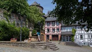 Ein gepflasterter Platz mit historischem Fachwerkhaus und altem Steinturm im Hintergrund, davor entnimmt eine Person Wasser aus einem steinernen Brunnen. Umgeben von Bäumen, Steinmauer und Straßenlaternen.