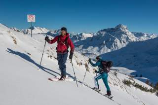 Zwei Skitourengeher steigen bei sonnigem Wetter einen schneebedeckten Berghang in den Alpen hinauf, im Hintergrund sind schneebedeckte Berggipfel zu sehen. Daneben steht ein Schild mit dem Hinweis „Stopp!“.