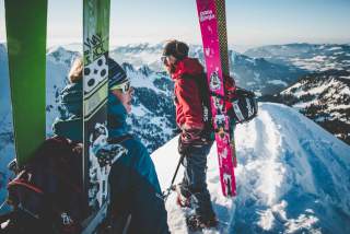 Zwei Menschen mit Ski an den Rucksäcken auf schneebedecktem Grat