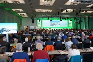 Großer Konferenzraum mit vielen Teilnehmenden, die einem Vortrag vor einer großen Bühne mit grüner Hintergrundbeleuchtung und mehreren Bildschirmen folgen.