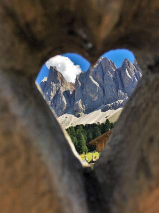 Durch ein herzförmiges Guckloch in einem Holzzaun sind steile Felsformationen und eine Hütte zu sehen.