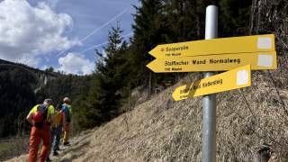 Drei Wanderer gehen auf einem schmalen Pfad bergauf, daneben steht ein gelber Wegweiser mit Richtungen zur Seaperalm, Stafflacher Wand Normalweg und Stafflacher Wand Klettersteig. Im Hintergrund sind Bäume, Himmel mit Wolken und eine Brücke zu sehen.