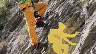Ein Klettersteiggeher mit Helm, Sonnenbrille und Rucksack klettert an einem Klettersteig an einer steilen Felswand, daneben eine gelbe Metallfigur eines Kletterers mit der Aufschrift „Peter Kofler Klettersteig“.