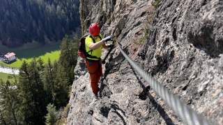 Eine Person mit Helm und Kletterausrüstung klettert an einem Drahtseil entlang einer steilen Felswand, im Hintergrund Wald und Wiese.