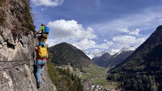 Zwei Menschen mit Klettersteigausrüstung erklimmen einen Felsen, im Hintergrund ein Tal mit Dorf und schneebedeckten Bergen.