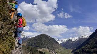 Zwei Personen in einem Klettersteig. Im Hintergrund der Talboden und schneebedeckte Berge.
