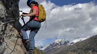 Eine Person mit Kletterausrüstung und Helm erklimmt eine Felswand, im Hintergrund schneebedeckte Berge und bewaldete Hänge unter wolkigem Himmel.
