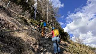 Zwei Personen mit Klettersteigausrüstung steigen einen steilen, felsigen Hang hinauf und halten sich an einem Stahlseil fest; im Hintergrund Bäume und blauer Himmel mit Wolken.