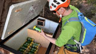 Ein Klettersteiggeher mit Helm und Rucksack nimmt eine Getränkedose aus einer geöffneten Holztruhe im Wald.