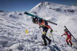 Zwei Athleten laufen mit Ski auf dem Rücken Berg rauf