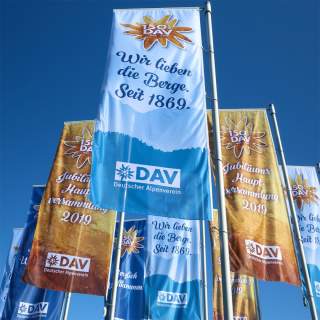 Mehrere Fahnen wehen im Wind, die den 150. Jahrestag (1869-2019) des Deutschen Alpenvereins (DAV) mit den Worten „Wir lieben die Berge. Seit 1869.“ und „Jubiläums Hauptversammlung 2019“ feiern.