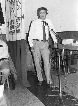 Schwarz-Weiß-Foto von Josef Klenner. Er steht vor einem Mikrofon in einem Sitzungssaal, neben ihm hängt ein Stoffbanner mit Edelweiß.