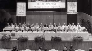 Hauptversammlung des DAV in Ingolstadt 1992. Ein altes Schwarz-Weiß-Bild zeigt den Sitzungssaal. Auf einem Podium sind zwei Reihen mit Menschen besetzt, 30 Personen sind auf der Bühne zu sehen. Hinter ihnen hängen Banner mit der Schrift "DAV-Hauptversammlung 28.-30. Mai 1992, 110 Jahre Sektion Ingolstadt" sowie Banner mit Edelweiß und der Schrift "Sektion Ingolstadt" und "Sektion Kaiserslautern".