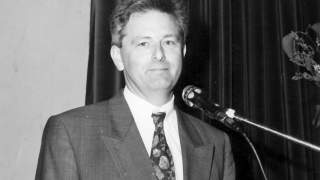 Josef Klenner 1992. Schwarz-Weiß-Foto eines Mannes im Anzug. Er blickt in die Kamera, vor ihm steht ein Mikrofon, hinter ihm ist ein dunkler Vorhang zu sehen.