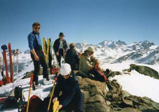 Ein altes Farbbild zeigt eine Gruppe von fünf Personen in den Bergen. Die Landschaft ist zum Teil verschneit, die Menschen sitzen auf Felsen. Sie haben Skiausrüstung dabei.