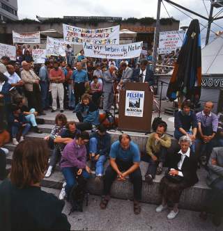 Altes Farbfoto von einer Demonstration, Menschen mit Bannern und bunter Schrift, vorne im Bild sitzen mehrere Leute auf Betonstufen, in der Mitte ein Rednerpult, an dem Josef Klenner steht.
