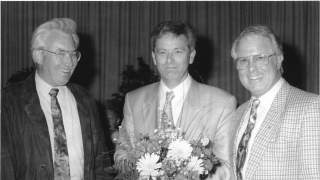 Vorstand des DAV. Schwarz-Weiß-Bild zeigt drei Männer vor einem dunklen Vorhang. Sie lächeln in die Kamera. Josef Klenner in der Mitte hält einen Blumenstrauß.