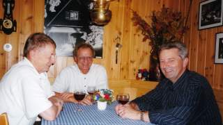 Vorstand des DAV 2000. Altes Farbfoto zeigt drei Männer. Sie sitzen an einem Tisch mit blau-weiß karierter Tischdecke, vor jedem steht ein Glas Rotwein. Der Raum ist holzgetäfelt, an der Wand hängen Bilder und Plakate, auf der Rückenlehne der Eckbank stehen Trockenblumen.