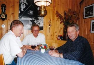 Vorstand des DAV 2000. Altes Farbfoto zeigt drei Männer. Sie sitzen an einem Tisch mit blau-weiß karierter Tischdecke, vor jedem steht ein Glas Rotwein. Der Raum ist holzgetäfelt, an der Wand hängen Bilder und Plakate, auf der Rückenlehne der Eckbank stehen Trockenblumen.