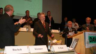 Abschied Josef Klenner Hauptversammlung. Auf einer Bühne stehen Tische und ein Rednerpult. Menschen klatschen stehend, die Blicke sind auf Josef Klenner in der Bildmitte gerichtet, er lacht.