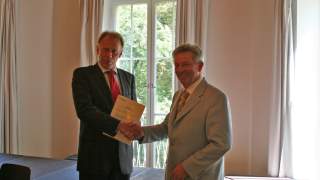 Jürgen Trittin und Josef Klenner, Anerkennung des DAV als Naturschutzverband. Zwei Männer in Anzügen schütteln sich die Hand und blicken in die Kamera. Einer hält Papiere in der Hand. Im Hintergrund helle Vorhänge und Wände, durch ein bodentiefes Fenster sind Bäume zu sehen.