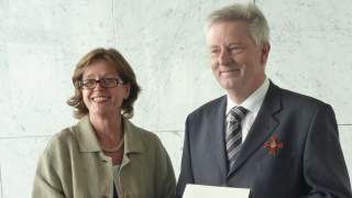 Ministerin übergibt Bundesverdienstkreuz an Josef Klenner. Zwei Personen stehen vor einer weißen Marmorwand und lächeln in die Kamera. Die Frau links hält eine dunkelblaue Schatulle, der Mann rechts eine weiße Mappe, auf beiden ist der Bundesadler in Gold gedruckt.