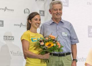 Jule Wurm und Josef Klenner 2015. Vor einer weißen Wand mit grauen Logos stehen eine Frau und ein Mann, beide Lachen. Die Frau hält einen Blumenstrauß.