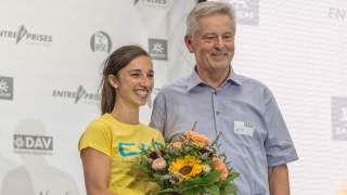 Jule Wurm und Josef Klenner 2015. Vor einer weißen Wand mit grauen Logos stehen eine Frau und ein Mann, beide Lachen. Die Frau hält einen Blumenstrauß.
