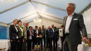 Fassanstich in Bierzelt. Mehrere Personen stehen in einem kleinen Bierzelt. Rechts im Bild: Josef Klenner deutet auf den Zapfhahn im Bierfass, in der rechten Hand hält er ein Bierglas, das fast vollständig mit Schaum gefüllt ist.