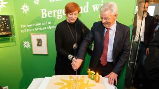 Melanie Grimm und Josef Klenner bei der Jubiläumsfeier 2019. Sie halten gemeinsam ein Messer mit dem sie einen flachen, rechteckigen Kuchen fast durchgeschnitten haben. Der Kuchen ist mit hellem Fondant gedeckt, auf einem goldenen Edelweiß steht "150 Jahre DAV, 1869-2019", im rechten Eck brennen drei Kerzen in Form einer "150".